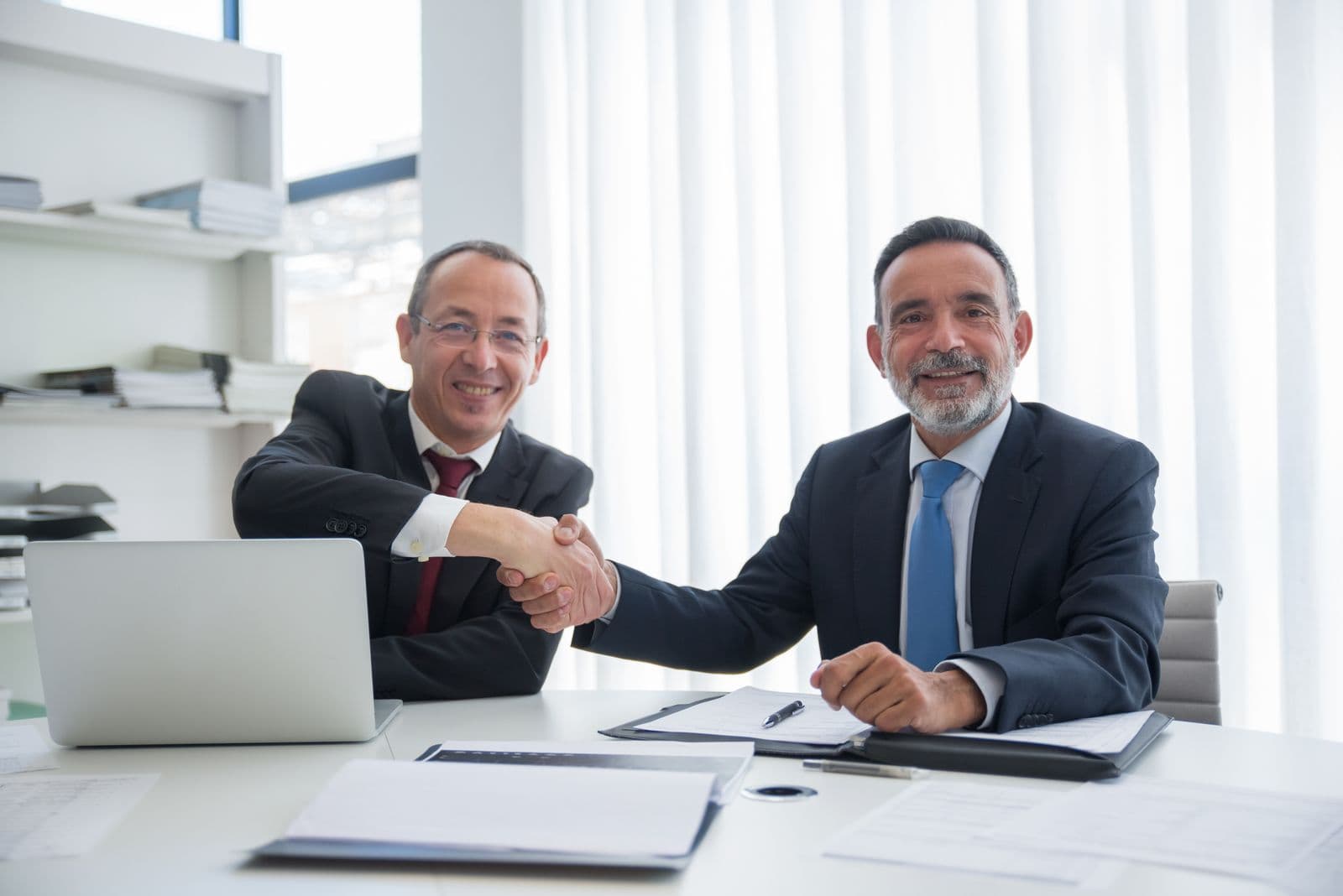Two men in suits shaking hands over paperwork at an office desk
